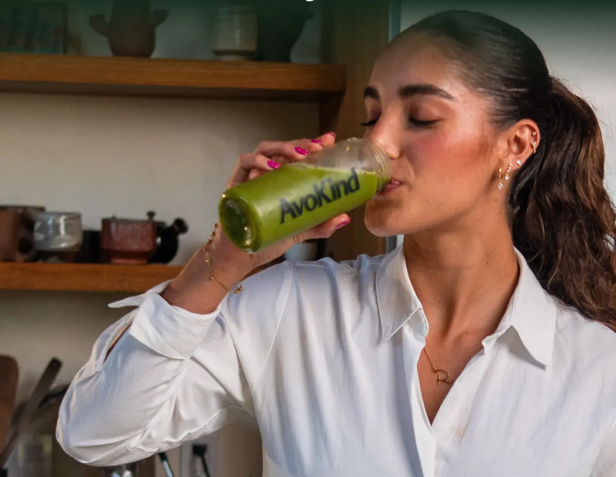 A woman drinking a green smoothie from an AvoKind bottle in a kitchen